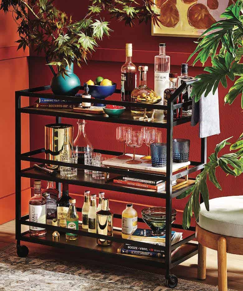 Stylish bar cart with liquor bottles, glassware, books, and decorative items against a red wall