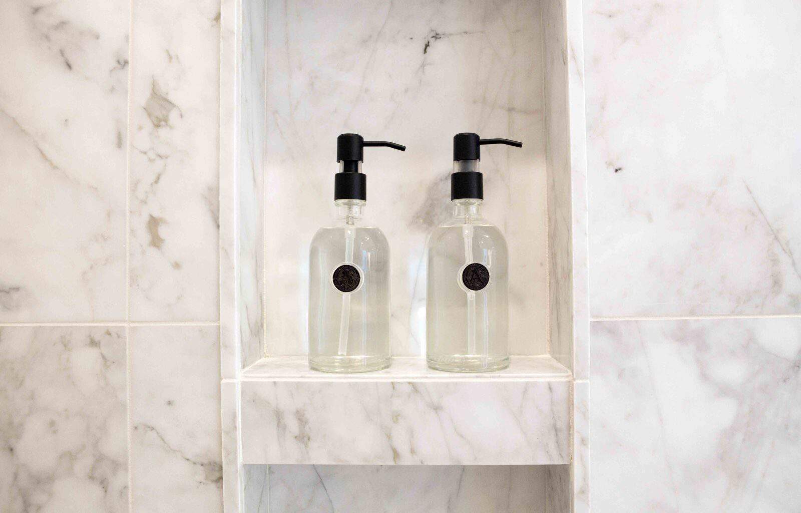 Two glass soap dispensers with black pumps placed on a marble shelf