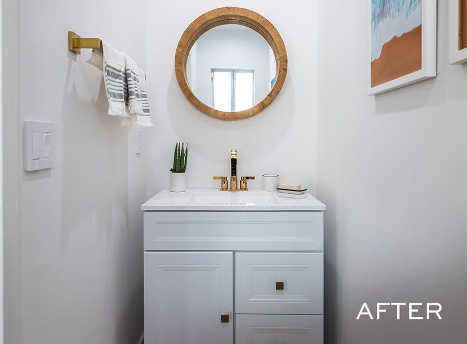 Small bathroom vanity area with a white cabinet, gold faucet, and a round wooden mirror