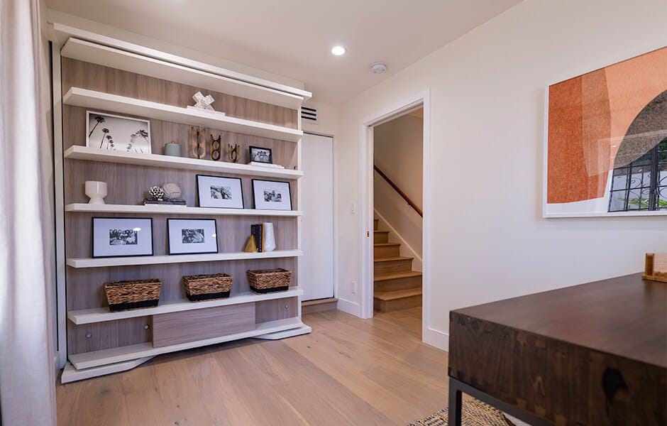 Hallway with built-in bookshelf, framed photos, vases, baskets, wooden floor, staircase, door, and abstract painting