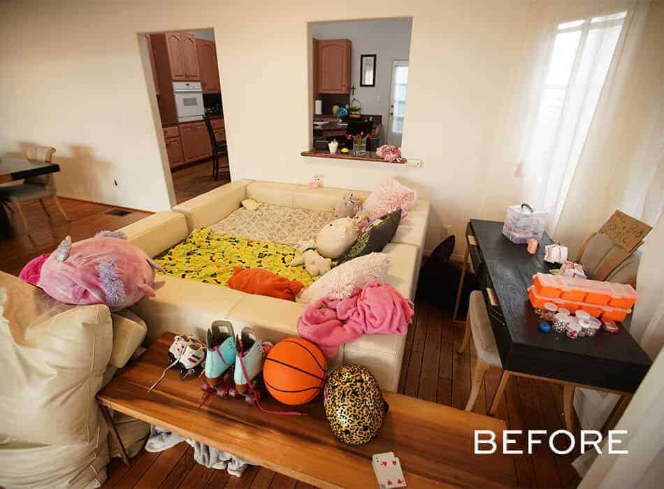 Cluttered living area with a sectional sofa, toys, and nearby craft table, with kitchen in the background