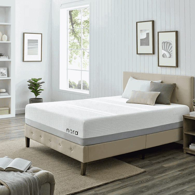 Beige upholstered bed with a white Nora mattress in a bright bedroom with framed artwork and a bookshelf