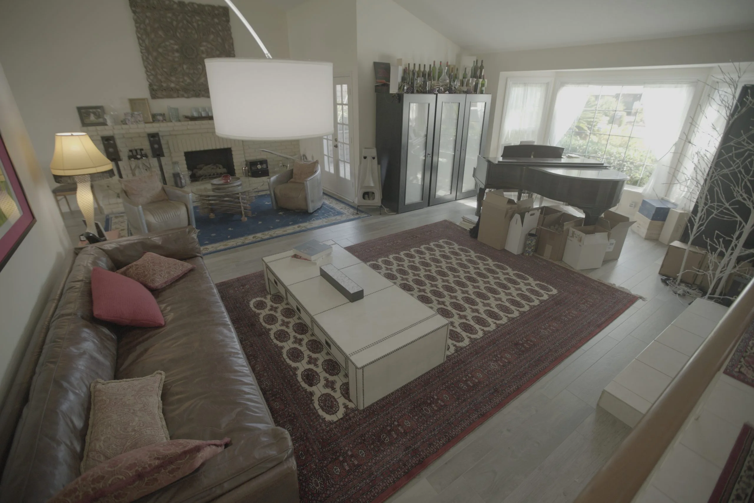 Cluttered living room with a leather sofa, patterned rugs, grand piano, and boxes near a large window