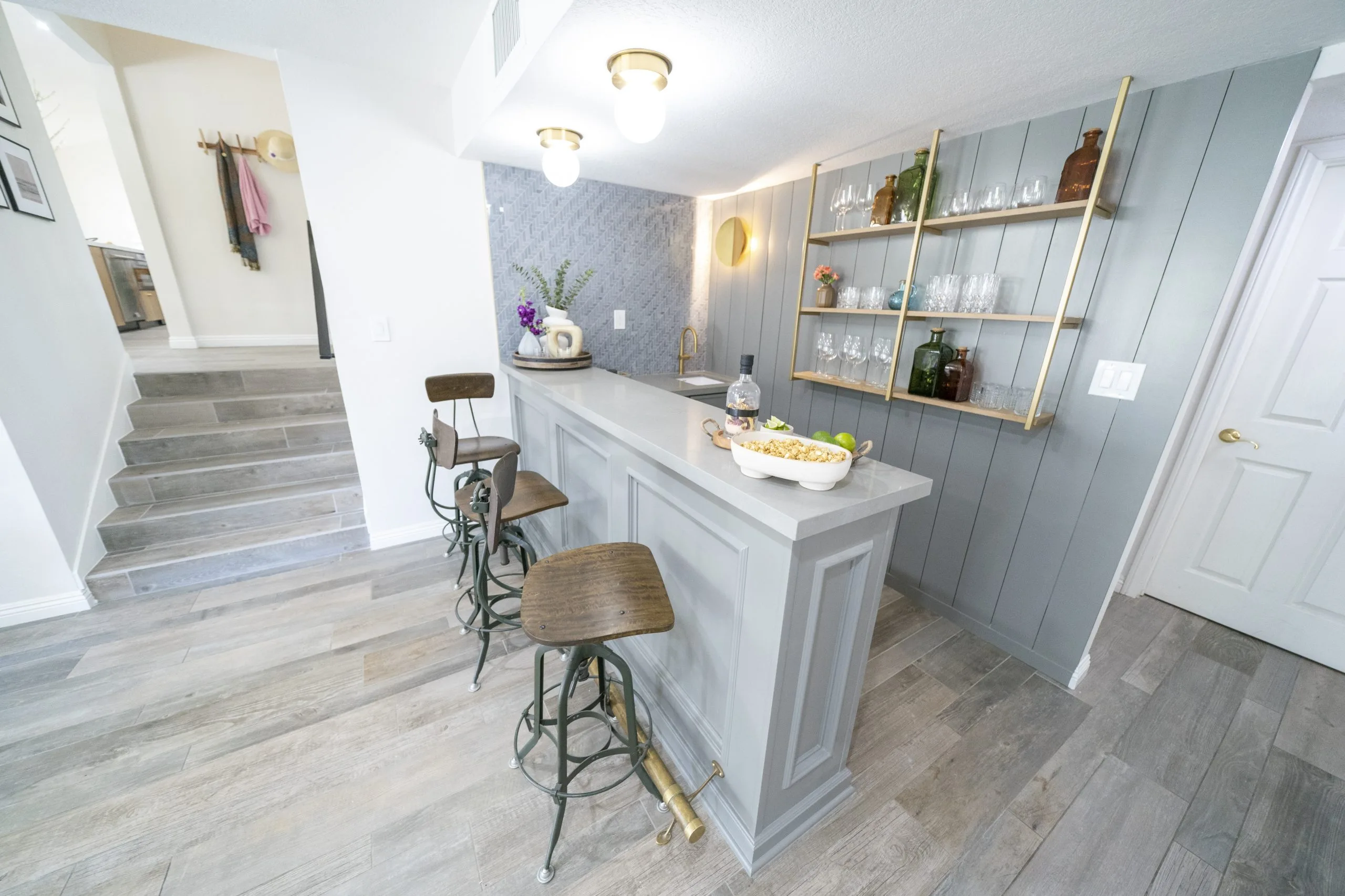 Modern home bar with gray paneling, open gold shelving, wooden bar stools, and decorative accents