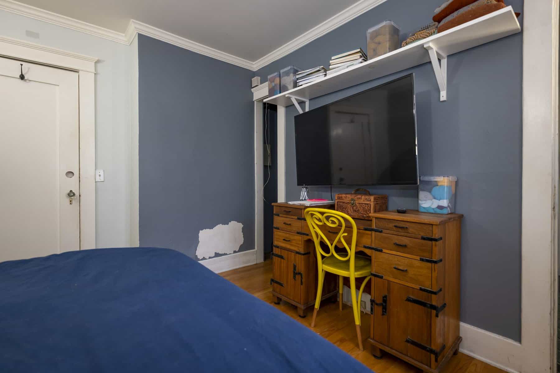 Bedroom with blue walls, wooden desk, yellow chair, and wall-mounted TV